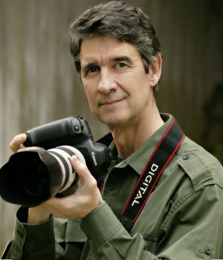 man with salt and pepper hair and army green jacket with camera looking at camera that is taking picture, photographer, photography classes, 