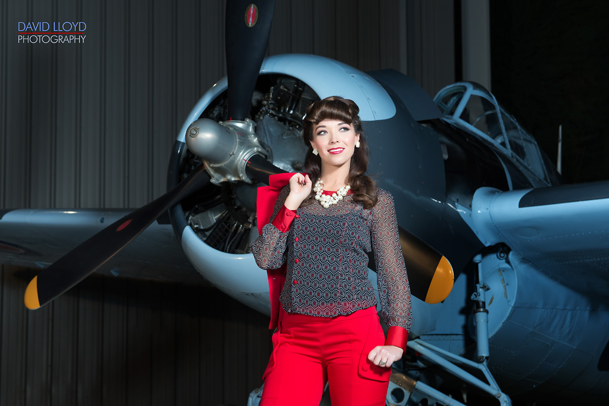 woman wearing red pantsuit with jacket thrown over her shoulder standing in a hanger in front of old military plane, fashion photography, david lloyd photography, glamour photography, 